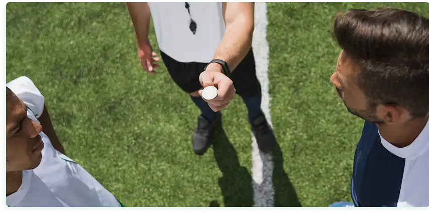 Op de afbeelding is een voetbalveld van bovenaf te zien. De scheidsrechter in een wit shirt staat in het midden van het veld en gooit een munt op voor aanvang van de wedstrijd, terwijl twee spelers van verschillende teams toekijken bij de toss. Bij de middenlijn ligt een voetbal en het zonlicht werpt scherpe schaduwen op het kunstgras.